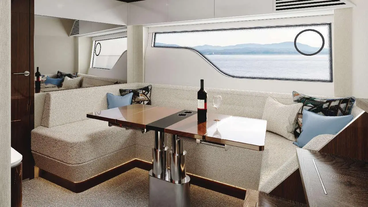 Modern yacht interior featuring a seating area, wooden dining table, wine bottle, and views of mountains through large windows.